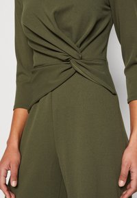 Femme portant un haut à manches longues et un pantalon vert olive avec un détail de nœud torsadé à la taille, mains détendues le long du corps.