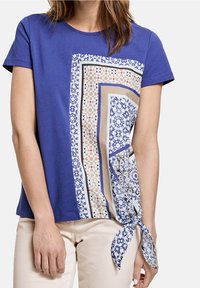 Woman wearing a short-sleeve blue top with a multi-patterned geometric scarf design on the front, tied in a knot at the hem.