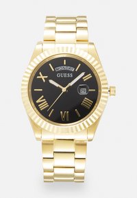 Montre en or avec un boîtier rond, un cadran noir, des index horaires en chiffres romains et un affichage jour/date. Terminée par un bracelet en maillons métalliques dorés.