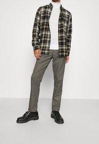 Camisa de franela en cuadro negro, amarillo y blanco; cuello de tortuga blanco; pantalones grises con patrón; zapatos negros con suela gruesa.