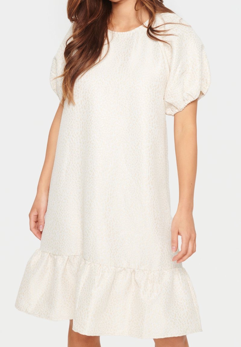 Femme portant une robe blanche ample à manches courtes bouffantes et un ourlet à volants, debout devant un fond uni.