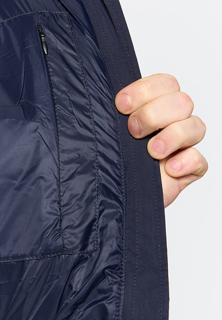 Intérieur de manteau rembourré bleu marine avec une texture lisse. Comprend une poche zippée sur le côté gauche et une main tenant le bord.