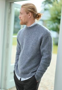 Pull en laine gris clair avec un col rond et des poignets côtelés, superposé sur une chemise à col à rayures blanches et bleues, associé à un pantalon sombre.