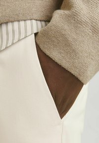 Beżowy sweter dzianinowy nałożony na jasną koszulę w paski, częściowo włożony w jasne spodnie z brązową kieszenią, o gładkich fakturach.