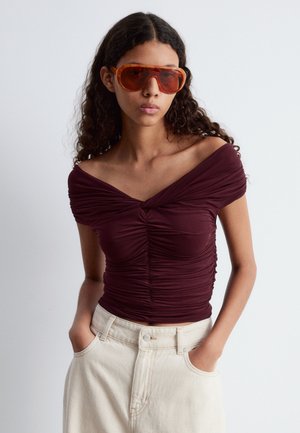 Bordeauxfarbene Off-Shoulder-Bluse mit Rüschen, figurbetontem Schnitt und Schlüsselloch-Ausschnitt; kombiniert mit hoch sitzenden beigen Jeans und übergroßen orangen Sonnenbrillen.