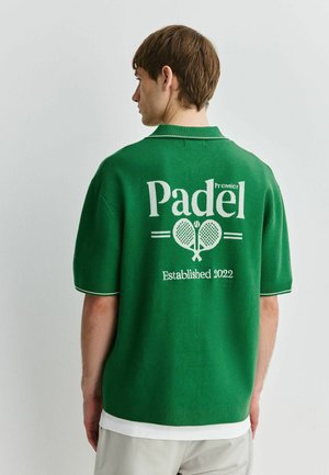 Tânăr cu spatele spre cameră poartă un tricou polo verde cu inscripția „Padel Premier Established 2022” și grafică cu rachete de padel încrucișate pe spate.