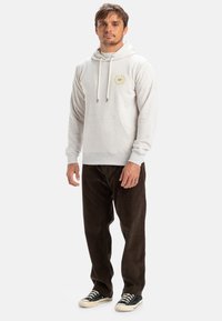 Lys grå hættetrøje med en frontlomme, justerbare snore og et logo på brystet, parret med mørkebrun kordfløjl bukser og sorte sneakers.