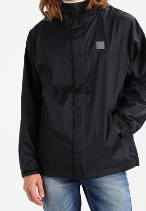 Zwarte windbreaker jas gemaakt van lichtgewicht, glanzende stof. Heeft een hoge kraag, elastische manchetten en een klein vierkant logo op de borst.