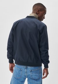 Marineblauwe bomberjack met een gladde textuur, geribbelde manchetten en een contrasterende lichte kraag. Draagt over lichtblauwe jeans, met een ontspannen pasvorm.