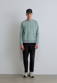 Világoszöld pulóver dombornyomott "BOSS" logóval, fekete szűkített nadrág, fehér sportcipő, fekete sapka logóval kiegészítve.