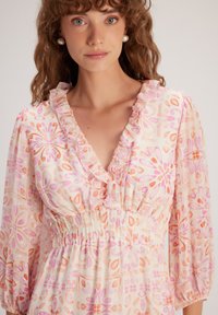 Blouse à motif floral en tissu doux et léger, avec un col froncé, une taille élastique et des manches bouffantes. Les couleurs comprennent le rose et l'orange.