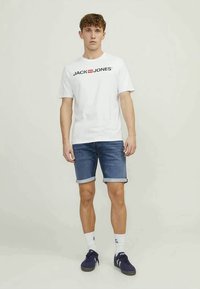 T-shirt branca com o logótipo "JACK & JONES", calções de ganga azuis com as bainhas dobradas, meias brancas e ténis escuros. Conjunto casual simples.
