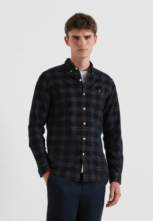Camisa de cuadro oscuro con botones en tonos de azul marino, marrón y crema, con cuello puntiagudo y logo en el bolsillo delantero. Llevada sobre una camiseta blanca.