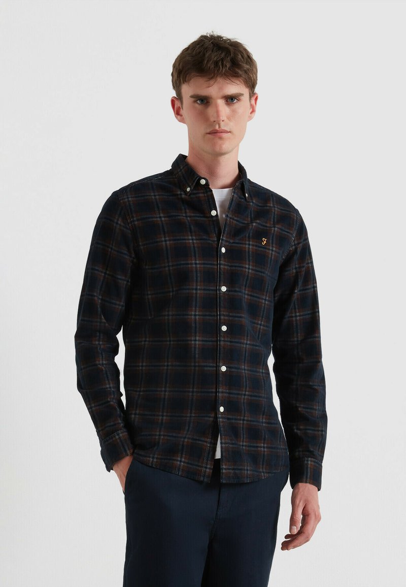 Camisa de cuadro oscuro con botones en tonos de azul marino, marrón y crema, con cuello puntiagudo y logo en el bolsillo delantero. Llevada sobre una camiseta blanca.