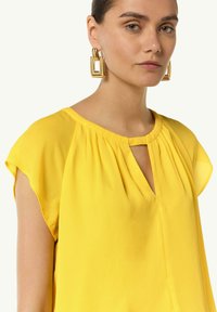 Żółta bluzka krótko rękawowa z dekoltem w kształcie łezki, wykonana z lekkiego materiału. Posiada delikatne marszczenie wzdłuż ramion i dekoltu.