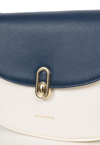 Borsa a tracolla con parte superiore testurizzata blu navy e parte inferiore liscia bianca. Presenta una chiusura dorata e il logo "HEXAGONA" impresso sul davanti.