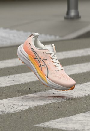 Chaussure de running rose clair avec des accents orange flottant au-dessus d'un passage piéton sur une route en asphalte.