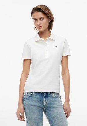 Žena s krátkymi vlasmi, oblečená v bielej polokošeli s krátkym rukávom a svetlomodrých džínsoch, stojaca na jednofarebnom pozadí.