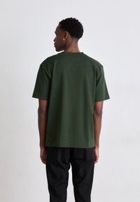 T-shirt en coton vert foncé avec un col rond et des manches courtes, présentant une coupe décontractée et sans motifs ni graphismes visibles.