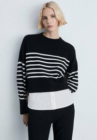 Schwarz-weiß gestreifter Pullover mit rundem Halsausschnitt und langen Ärmeln, über einem weißen Hemd mit Kragen getragen, kombiniert mit einer schwarzen Hose.