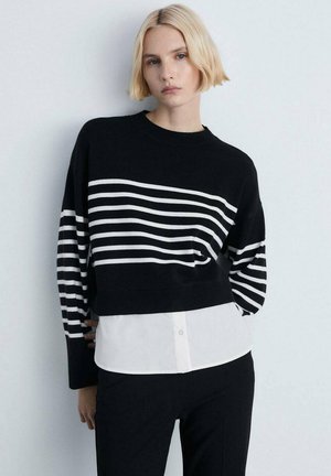 Pull rayé noir et blanc avec un col rond et des manches longues, superposé sur une chemise blanche à col, porté avec un pantalon noir.