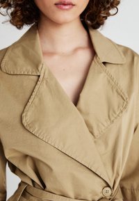 Beżowy trencz z dużymi klapami i dopasowaną talią. Posiada pojedyncze zapięcie na guzik oraz gładki, lekko teksturowany materiał.