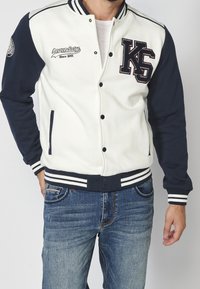 Veste de varsity blanche et marine avec un col en tricot, dotée de logos brodés, de boutons-pression et de poches latérales. Tissu texturé avec des rayures.