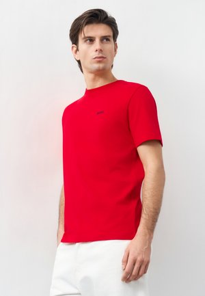 Άνδρας που φοράει φωτεινό κόκκινο κοντομάνικο μπλουζάκι με το λογότυπο "BOSS", συνδυασμένο με λευκό παντελόνι, στέκεται μπροστά σε απλό ανοιχτόχρωμο φόντο.