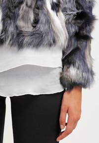Personne portant une veste en fausse fourrure grise et blanche par-dessus un haut blanc superposé avec un pantalon noir, montrant une main détendue sur le côté.