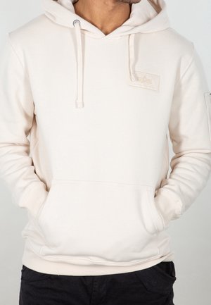 Mężczyzna w prostym kremowym kapturze z przednią kieszenią i subtelną naszywką z logo, ręce w kieszeniach, ubrany w czarne spodnie na neutralnym tle.