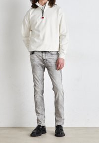 Biały pulower z półzamkiem, szare jeansy w stylu distressed, czarne mokasyny. Casualowy design z teksturowanym wykończeniem, z czerwonym akcentem na zamku.