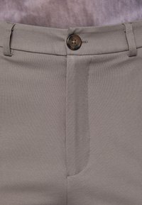 Grijze stoffen broek met een gladde textuur, platte voorkant en een enkele bruine knoopsluiting bij de tailleband. Stiksels zichtbaar langs de naden.