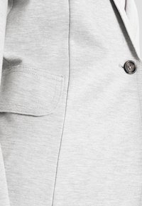 Ljusgrå jacka i texturerad tyg med en knappknäppning och en ficka på vänster sida.