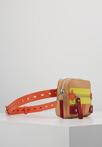 Kompakt crossbody-väska i beige läder med neongula och orange detaljer. Har ett dragkedjestängning, dekorativ hårdvara och justerbar rem.