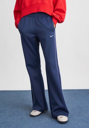 Persona che indossa pantaloni sportivi Nike blu navy con strisce laterali bianche, una felpa rossa e scarpe slip-on Nike di colore chiaro.