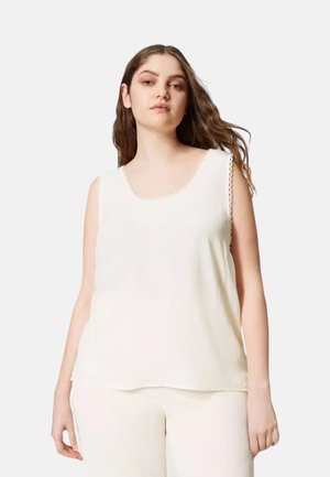 Donna con lunghi capelli castani, indossa un top bianco senza maniche e pantaloni bianchi, in piedi davanti a uno sfondo chiaro e uniforme.