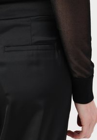 Crna majica dugih rukava od prozirnog materijala s rebrastim manžetama, uparena s elegantnim satenastim crnim hlačama koje imaju diskretnu stražnju džep.
