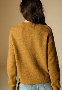 Senfgelbes Strickpullover mit rundem Halsausschnitt, langen Ärmeln und strukturiertem Finish. Kombiniert mit hellblauen Jeans.