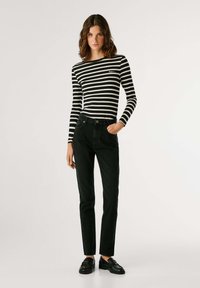 Melnas džinsa bikses, kas apvienotas ar pieguļošu melnbaltu svītrainu garo piedurknju topu, ar mazu logotipu uz krūšu daļas. Modelis pozē, ieliekot vienu roku kabatā.