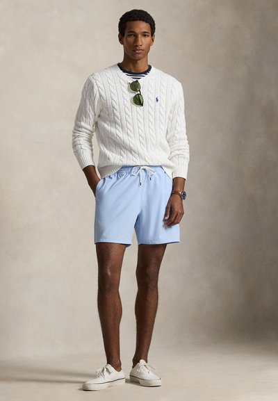 Άσπρο πουλόβερ πλεκτό με καλώδιο, μπλε σορτς και άσπρα αθλητικά παπούτσια. Το μοντέλο στέκεται με τα χέρια στις τσέπες. Αξεσουάρ περιλαμβάνουν πράσινα γυαλιά ηλίου.