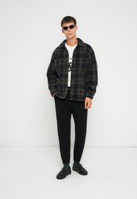 Jachetă din flanel cu carouri în albastru închis și bej, tricou grafic alb, pantaloni jogger negri și adidași închiși la culoare, completate cu ochelari de soare.