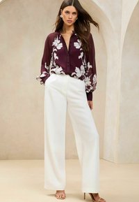 Maroon floral blouse with button-down front, long sleeves, paired with white wide-leg trousers and beige high-heeled sandals.