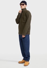 Fleece pullover in olive green with a half-zip collar, chest pocket, and textured finish. Paired with blue denim pants and tan shoes.