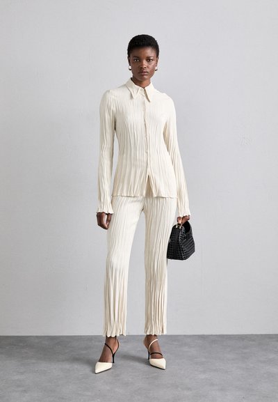 Cream-colored pleated shirt and pants set with long sleeves, collar, and ruffled cuffs; paired with black woven handbag and pointed heels.