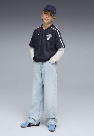 Niño con camiseta deportiva azul marino sobre mangas largas blancas, pantalones vaqueros de pierna ancha azul claro, gorra azul a juego y zapatillas, de pie con las manos en los bolsillos.
