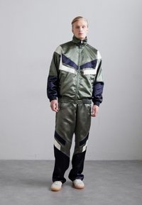 Survêtement satin vert avec une veste zippée aux accents blancs et bleu marine, associé à un pantalon fuselé assorti et des baskets blanches.