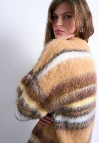 Jeune femme aux cheveux châtain clair portant un pull rayé et duveteux aux tons beige, blanc, marron et jaune, posant de profil devant un fond uni.