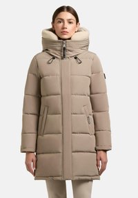 Donna che indossa un cappotto invernale imbottito beige con colletto alto foderato in felpa crema, chiusura frontale con zip e tasche laterali, in piedi davanti a uno sfondo bianco.