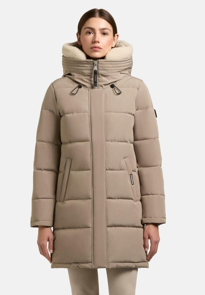Donna che indossa un cappotto invernale imbottito beige con colletto alto foderato in felpa crema, chiusura frontale con zip e tasche laterali, in piedi davanti a uno sfondo bianco.