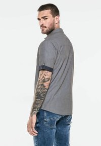 Grå kort ärmad skjorta med texturerad finish, mörk denimdetaljer på manschetterna, bärs med blå jeans, som visar en tatuerad arm.